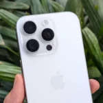 Điện thoại iPhone 16 Pro màu trắng cầm trên tay, phía sau là cây xanh, minh họa thiết kế và camera