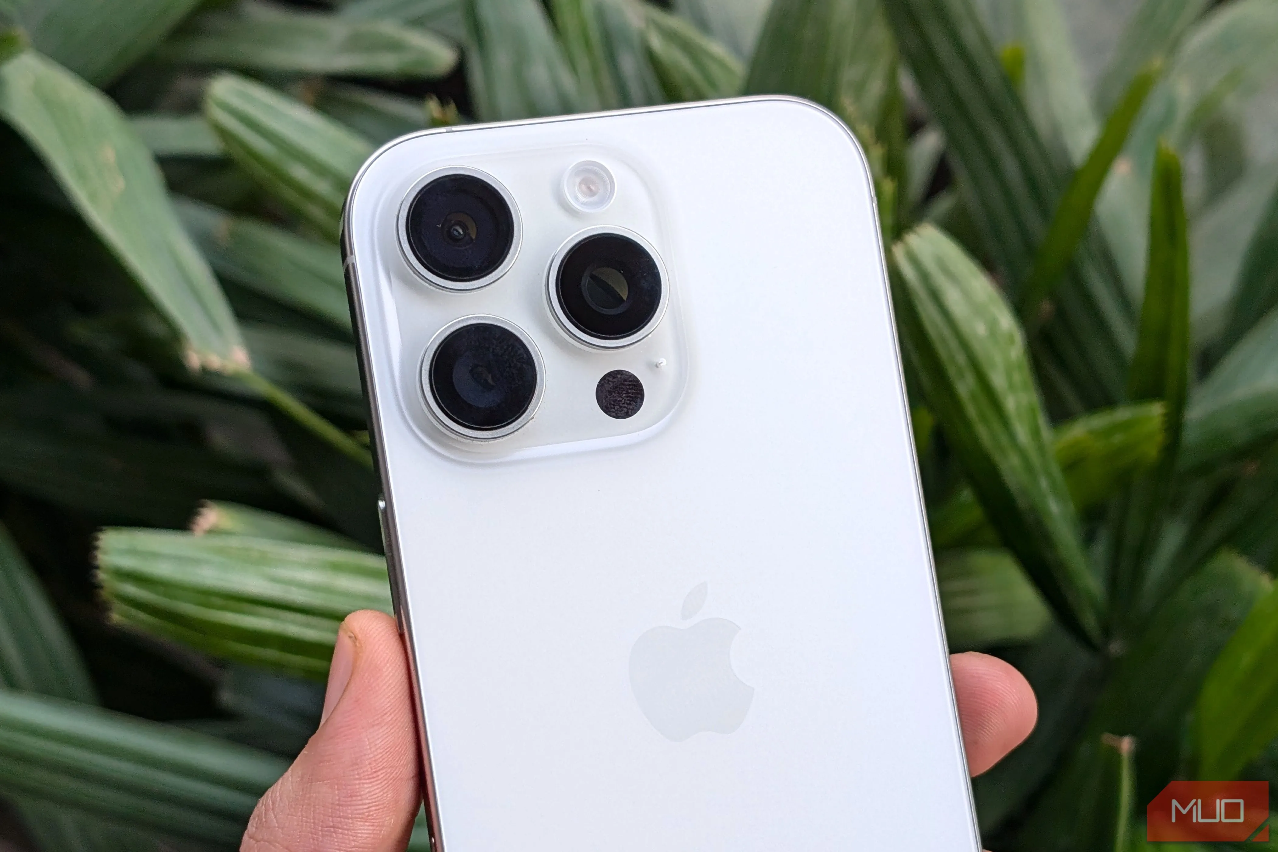 Điện thoại iPhone 16 Pro màu trắng cầm trên tay, phía sau là cây xanh, minh họa thiết kế và camera