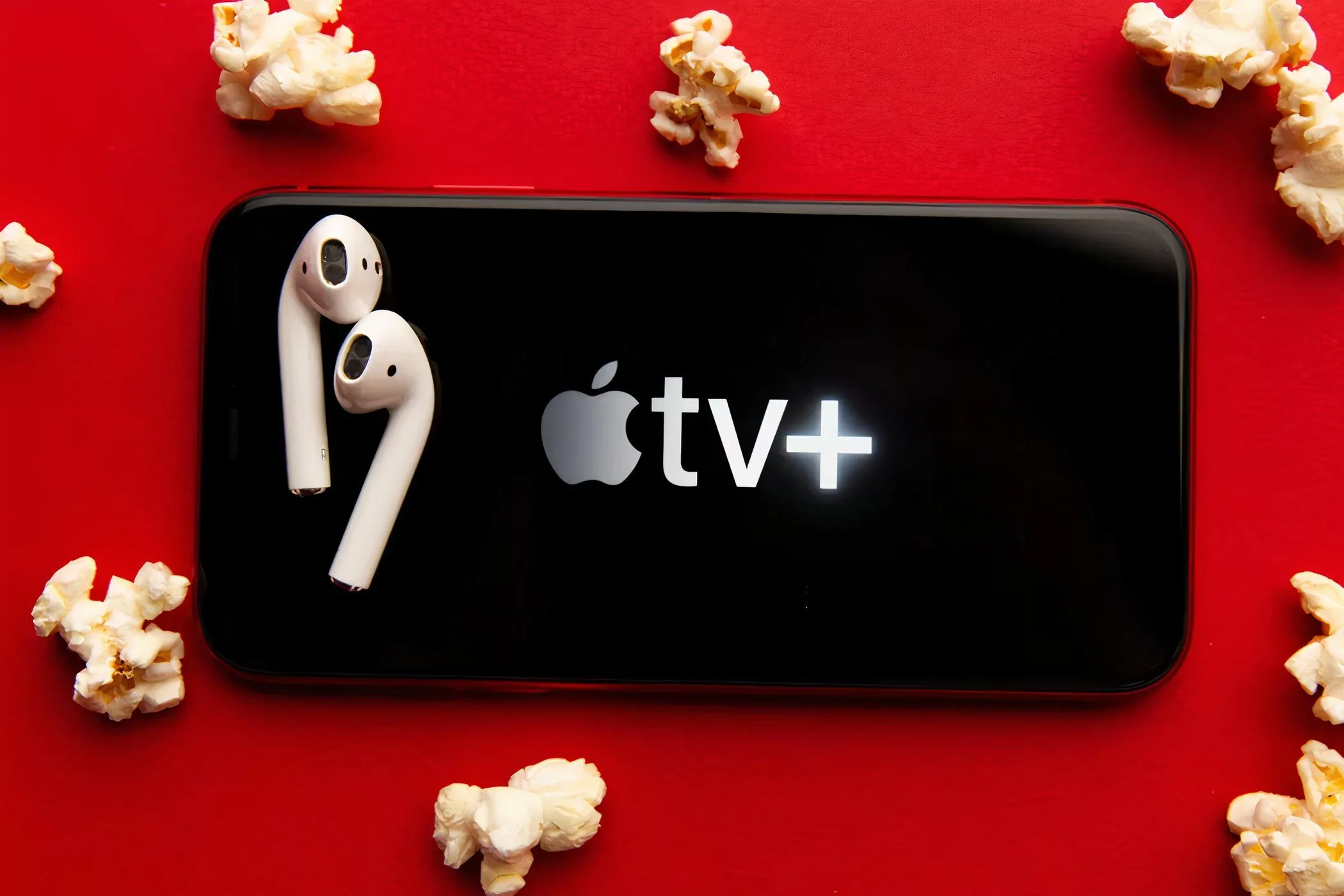 Màn hình điện thoại di động hiển thị logo Apple TV+, xung quanh là bỏng ngô và tai nghe, minh họa trải nghiệm xem phim thoải mái tại nhà.