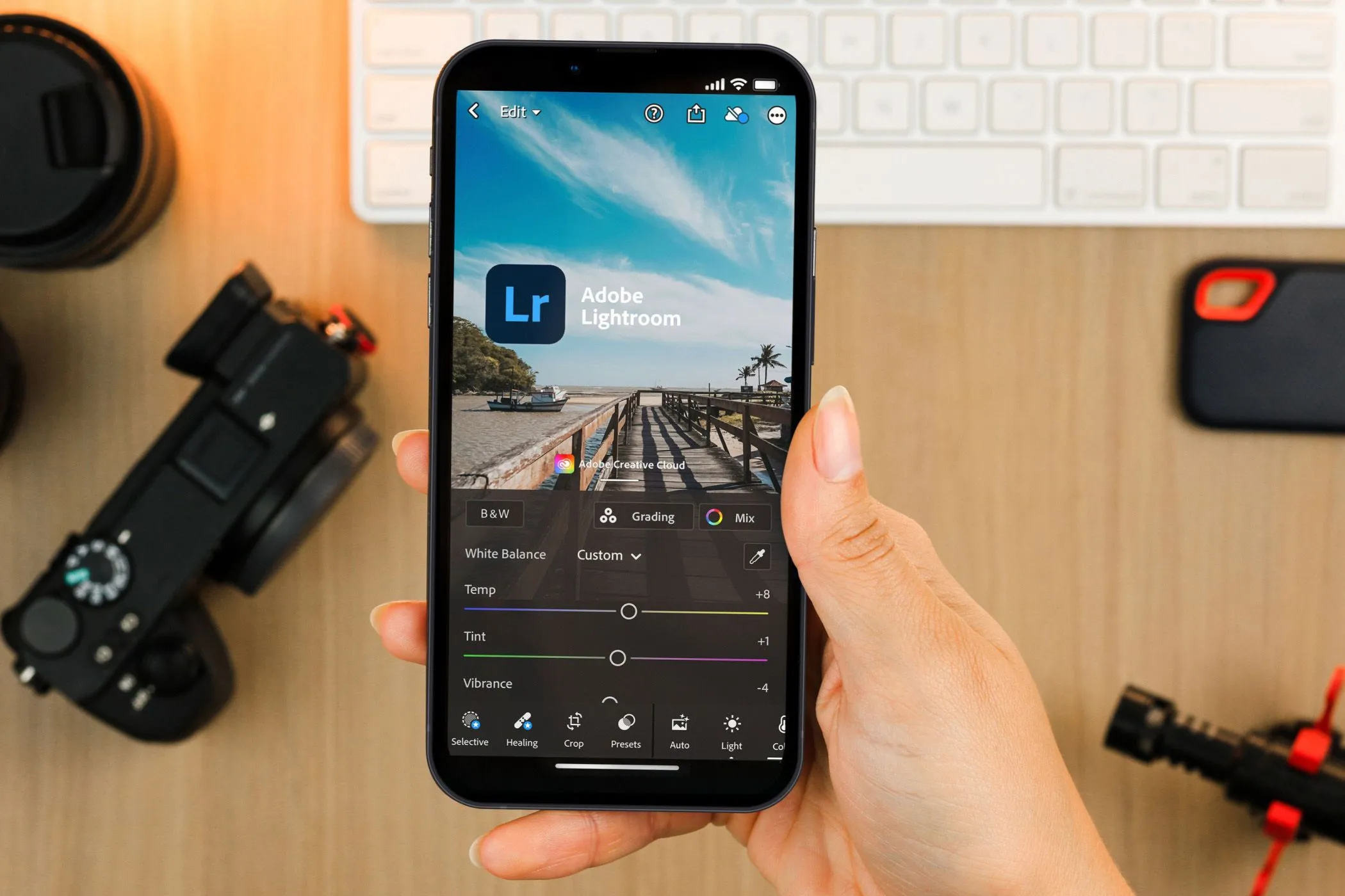 Một bàn tay đang cầm smartphone hiển thị giao diện ứng dụng Adobe Lightroom Mobile, với thiết bị chụp ảnh chuyên nghiệp ở hậu cảnh.